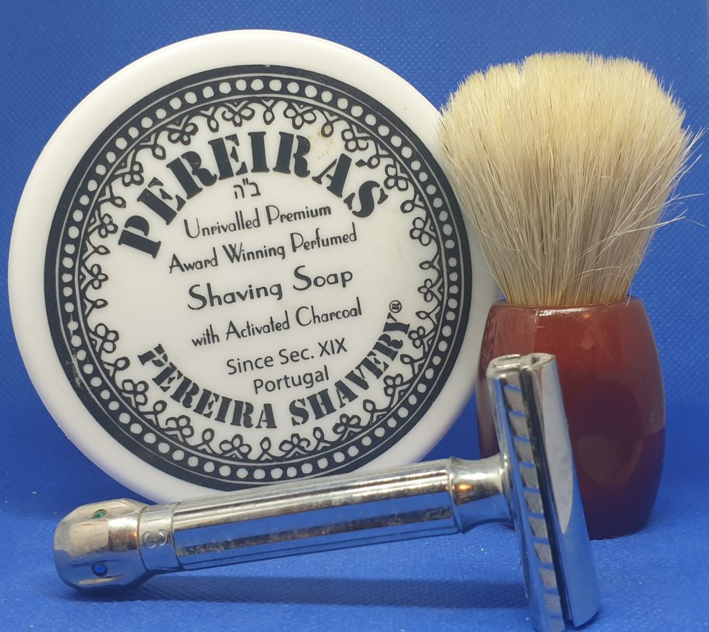 On a blue background; in front a shiny Mergress adjustable razor, behind it to the left a tub of Pereira shaving soap, and on the right a white horse hair shaving brush.