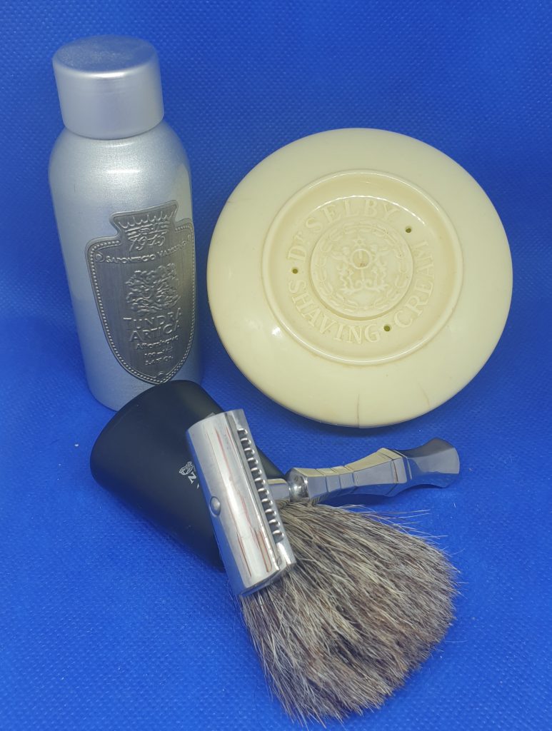 On a blue background; in front, a safety razor laying across a shaving brush.  Behind these, a metal Aftershave bottle and a plastic soap tub with embossed lid.
